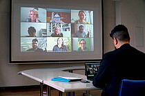 Auf dem Bild sieht man vorne etwas unscharf Klemens Ketelhut, der an einem Laptop sitzt. Scharf und in groß ist auf der Leindwand eine zoom-Sitzung zu sehen. An dieser nehmen das AW-ZIB sowie Franziska Brantner und Kathrin Rabus teil. Copyright: PH Heidelberg.