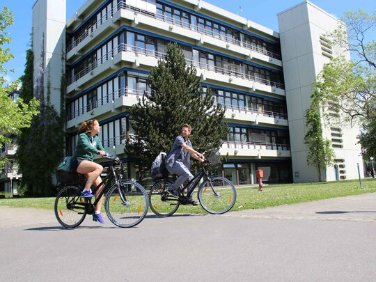 Eine junge Frau und ein junger Mann fahren auf Fahrrädern. Hier ihnen der Neubau der Pädagogischen Hochschule Heidelberg im Sommer.