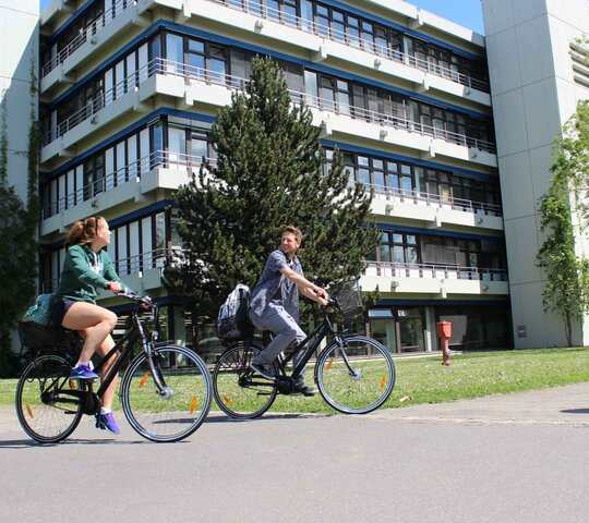 Eine junge Frau und ein junger Mann fahren auf Fahrrädern. Hier ihnen der Neubau der Pädagogischen Hochschule Heidelberg im Sommer.