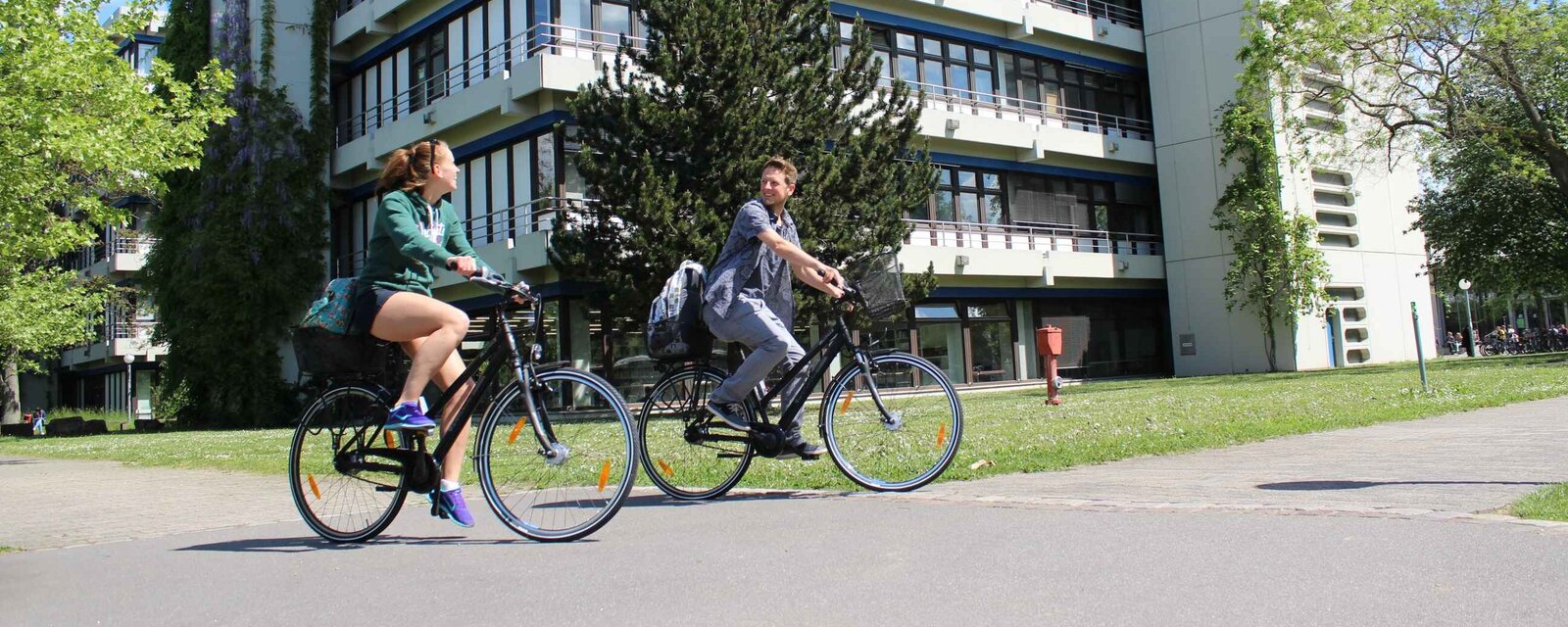 Eine junge Frau und ein junger Mann fahren auf Fahrrädern. Hier ihnen der Neubau der Pädagogischen Hochschule Heidelberg im Sommer.