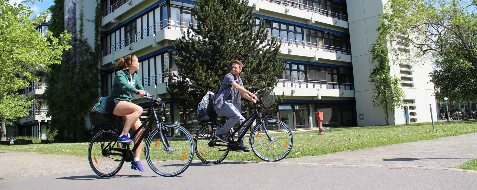 Eine junge Frau und ein junger Mann fahren auf Fahrrädern. Hier ihnen der Neubau der Pädagogischen Hochschule Heidelberg im Sommer.