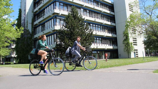 Eine junge Frau und ein junger Mann fahren auf Fahrrädern. Hier ihnen der Neubau der Pädagogischen Hochschule Heidelberg im Sommer.