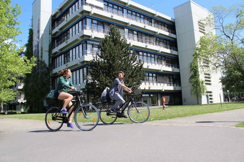 Eine junge Frau und ein junger Mann fahren auf Fahrrädern. Hier ihnen der Neubau der Pädagogischen Hochschule Heidelberg im Sommer.