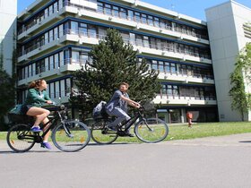 Eine junge Frau und ein junger Mann fahren auf Fahrrädern. Hier ihnen der Neubau der Pädagogischen Hochschule Heidelberg im Sommer.