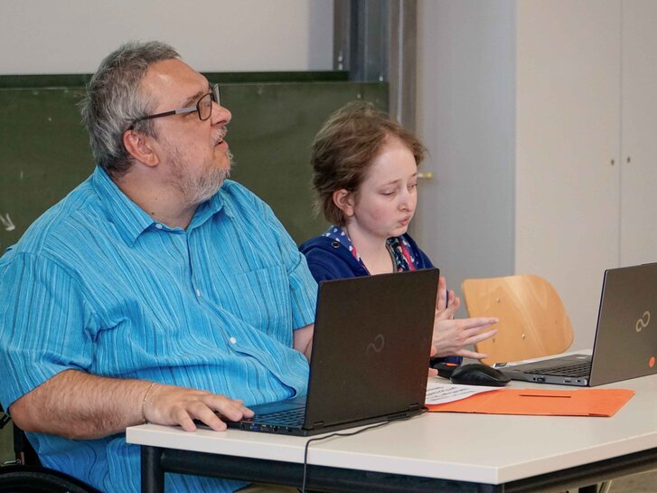 Helmuth Pflantzer und Louisa Kabbe sitzen an einem Tisch vor einer Tafel und bringen gemeinsam ein Bildungsangebot aus.