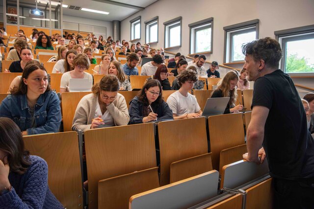 Ein Dozent steht vor einem Hörsaal, der voll besetzt ist mit Studierenden.