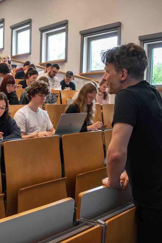 Ein Dozent steht vor einem Hörsaal, der voll besetzt ist mit Studierenden.