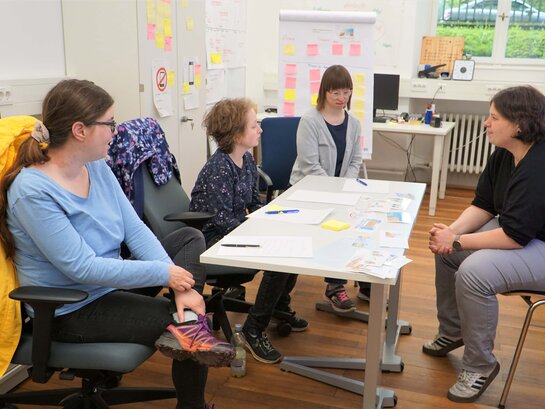Das Bild zeigt das Qualifizierungsbüro, in dem Susann Bensch, Louisa Kabbe, Anna Neff und Noemi Heister um einen Tisch herum sitzen. Im Hintergund steht ein FlipChart.