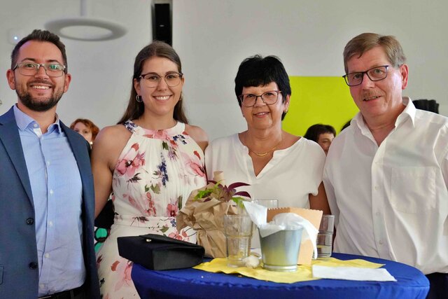 Zu sehen sind 4 Gäste ( 2 Männer außen, 2 Frauen dazwischen) an einem Stehtisch während des Sektempfangs, die alle in die Kamera schauen und lachen.