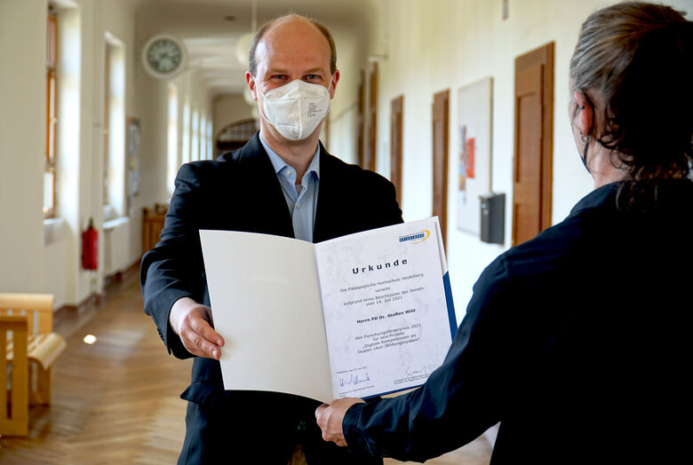 Das Bild zeigt primär Steffen Wild. Er schaut direkt in die Kamera und trägt Corona-bedingt einen Mund-Nase-Schutz. Vorne rechts sieht man Christian Spannagel von hinten. Er überreicht Wild die Urkunde. Das Foto wurde im Altbauflur der Hochschule aufgenommen. Copyright: PH Heidelberg