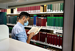 Das Symbolbild zeigt einen jungen Mann im Lesesaal der Hochschule. Er steht vor einem Regal und blättert in einem Buch. Er trägt Corona-bedingt einen Mund-Nase-Schutz.