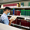 Das Symbolbild zeigt einen jungen Mann im Lesesaal der Hochschule. Er steht vor einem Regal und blättert in einem Buch. Er trägt Corona-bedingt einen Mund-Nase-Schutz.