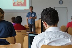 Das Foto zeigt Studierende in einem Hörsaal von hinten und vorne steht ein Professor.