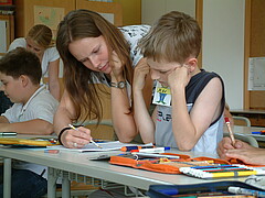 Das Foto zeigt einen Lehrenden der einem Kind am Tisch in der Schule bei einer Aufgabe hilft.