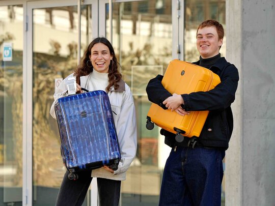 Zwei Studierende stehen am Campus und haben jeweils einen Reisekoffer in der Hand. Sie sind bereit für ihren Auslandsaufenthalt.