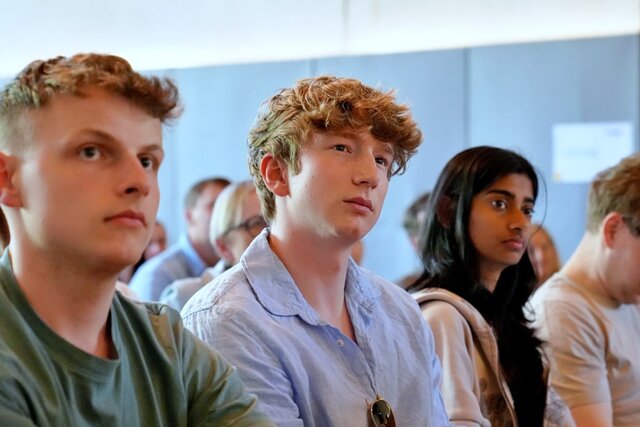 Zu sehen sind zwei Studenten sowie eine Studentin, sitzend, dem Vortrag zuhörend.