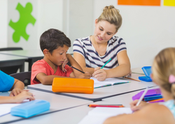 Lehrerin die neben ihrem Schüler am Tisch sitzt und ihm hilft.