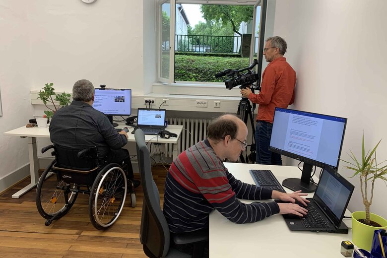 Das Foto zeigt zwei Bildungsfachkräfte im Büro. In der Ecke steht ein Kameramann.
