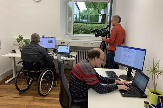 Das Foto zeigt zwei Bildungsfachkräfte im Büro. In der Ecke steht ein Kameramann.