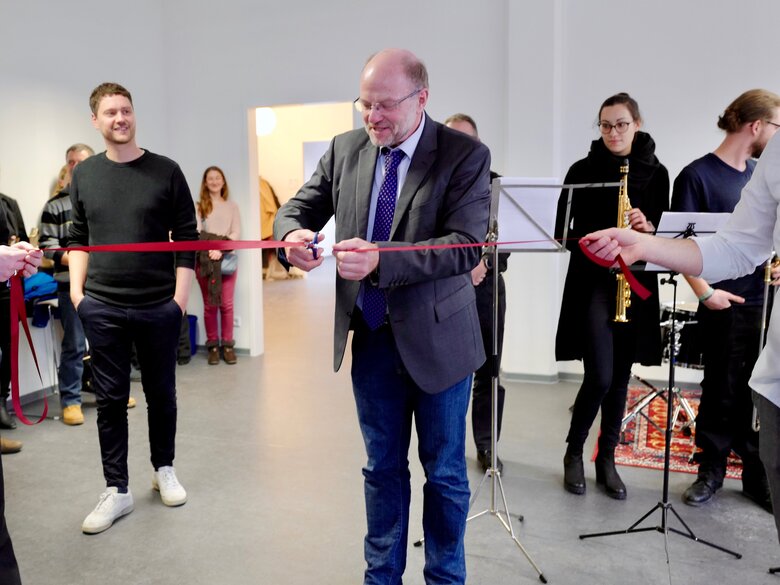 Zu sehen sind mehrere Personen bei einer Feierlichkeit. Eine Person durchschneidet ein rotes Band.