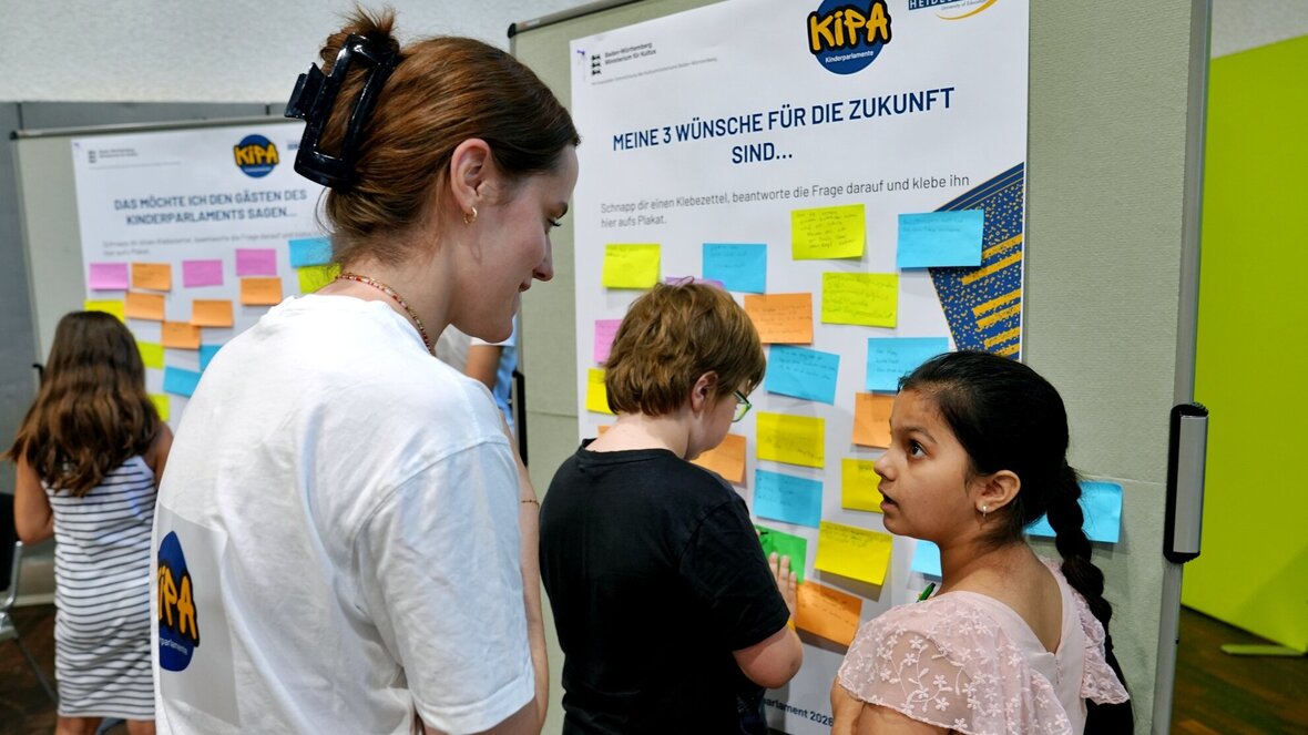 Eine Studentin spricht mit einer Grundschülerin. Im Hintergrund sieht man Kinder, die auf Plakaten des Kinderparlements ihre Stimme notieren. 