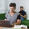 Auf dem Bild ist eine junge Frau am Laptop zu sehen. Hinter ihr sieht man unscharf einen Mann, der ein Buch liest. Copyright: Pädagogische Hochschule Heidelberg