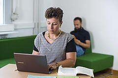 Auf dem Bild sieht man eine junge Frau. Sie arbeitet am Laptop. Im Hintergrund sieht man unscharf einen jungen Mann. Er stitz auf einer Coach und liest.