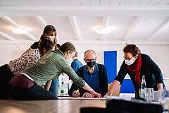 Das Bild zeigt mehrere KuLO-Teilnehmende, die gemeinsam Arbeiten. Sie sitzen oder stehen. Zwei Frauen zeigen auf ein Blatt, das auf einem Tisch zwischen den Personen liegt. Alle tragen Corona-bedingt einen Mund-Nase-Schutz.