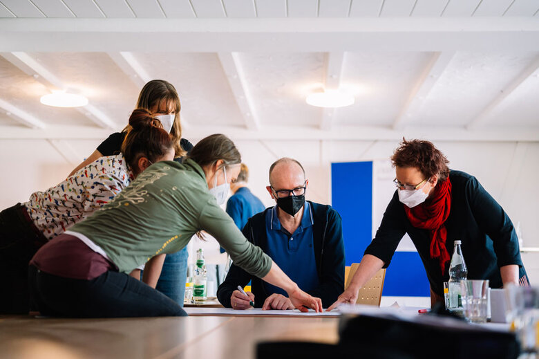 Das Bild zeigt mehrere KuLO-Teilnehmende, die gemeinsam Arbeiten. Sie sitzen oder stehen. Zwei Frauen zeigen auf ein Blatt, das auf einem Tisch zwischen den Personen liegt. Alle tragen Corona-bedingt einen Mund-Nase-Schutz.