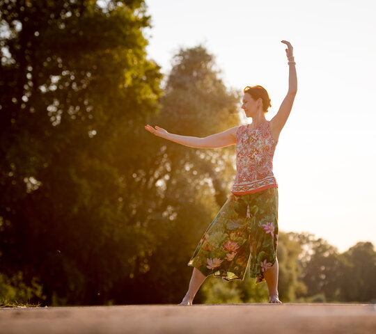 Frau steht im Sonnenuntergang auf Wiese und macht Qigong