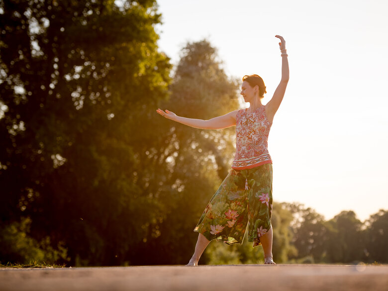Frau steht im Sonnenuntergang auf Wiese und macht Qigong