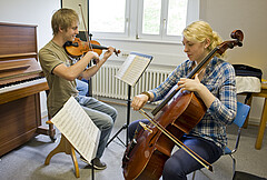 Das Bild zeigt links einen Studenten an der Geige und rechts eine Studentin, die Cello spielt. Die Aufnahme ist in den Überäumen der Hochschule entstanden.