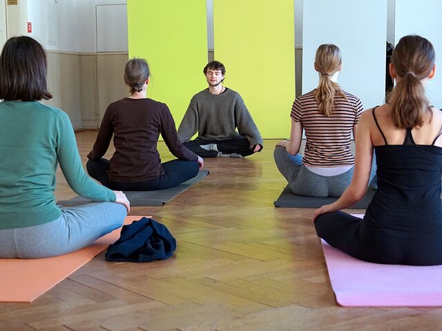 Yogalehrer und 4 Teilnehmerinnen sitzen im Schneidersitz auf Yogamatten