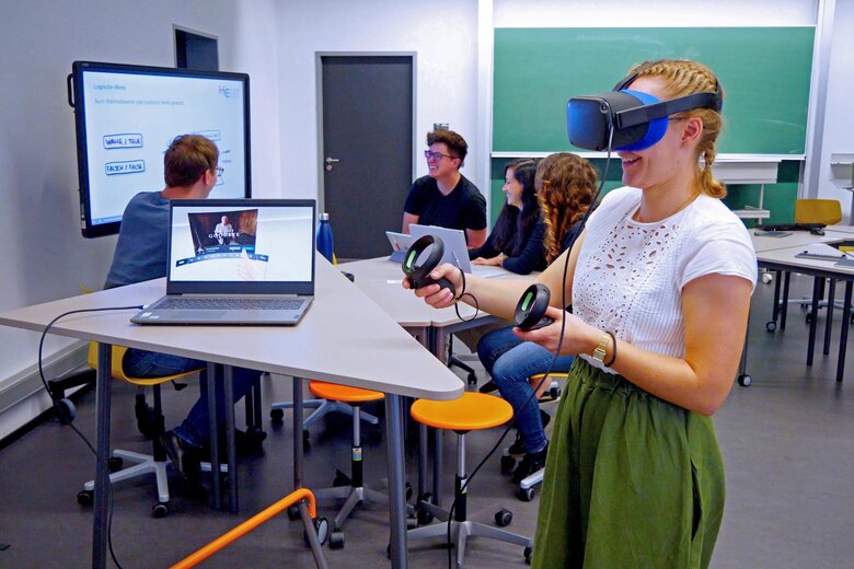 Ein Beispiel digitaler Lehre: Eine Studentin der Pädagogischen Hochschule Heidelberg trägt eine VR-Brille und hält die Controller in der Hand. Im Hintergrund sitzt eine Gruppe zusammen am Stehtisch und diskutiert vor einem Whiteboard.