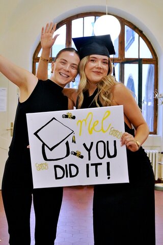 Zu sehen sind zwei junge Frauen, davon eine Absolvent:in mit Doktorhut & Schild auf dem steht "you did it" (rechts). 