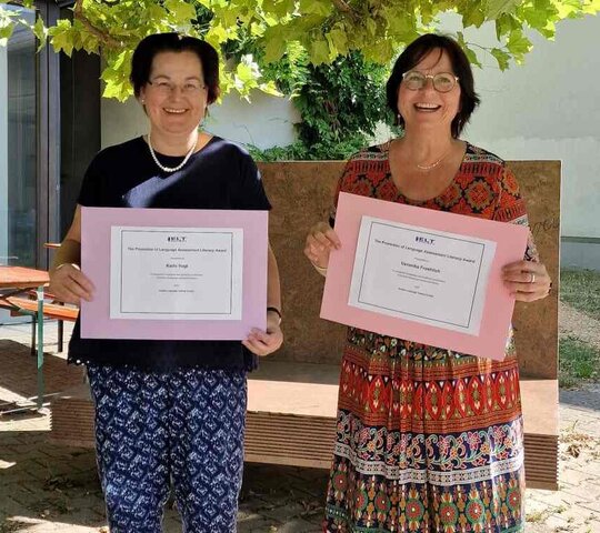 Karin Vogt und Veronika Fröhlich stehen im Innenhof des Neubaus und halten Zettel mit der Auszeichnung in die Kamera.