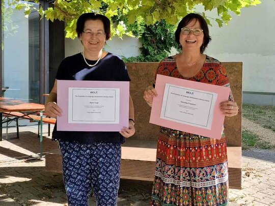 Karin Vogt und Veronika Fröhlich stehen im Innenhof des Neubaus und halten Zettel mit der Auszeichnung in die Kamera.