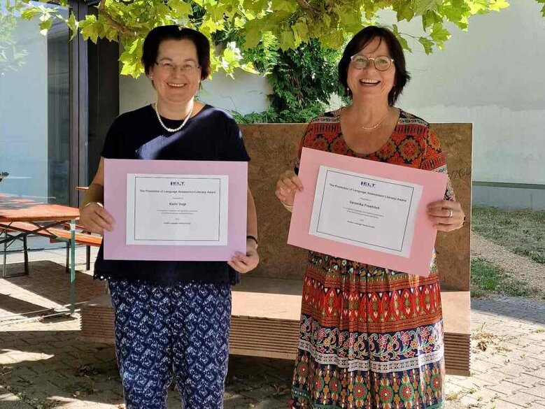 Karin Vogt und Veronika Fröhlich stehen im Innenhof des Neubaus und halten Zettel mit der Auszeichnung in die Kamera.