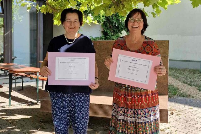Karin Vogt und Veronika Fröhlich im Innenhof des Neubaus der Pädagogischen Hochschule Heidelberg. Sie halten ihre Auszeichnung für Fremdsprachenunterricht in die Kamera, ein wichtiger Teil internationaler Lehre.