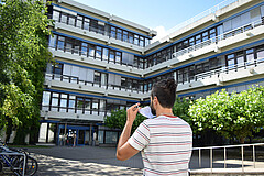 Das Symbolbild zeigt einen jungen Mann, der sich gerade eine Atemschutzmaske auszieht. Er steht vor dem Neubau der Hochschule und ist von hinten zu sehen. Copyright: Pädagogische Hochschule Heidelberg.