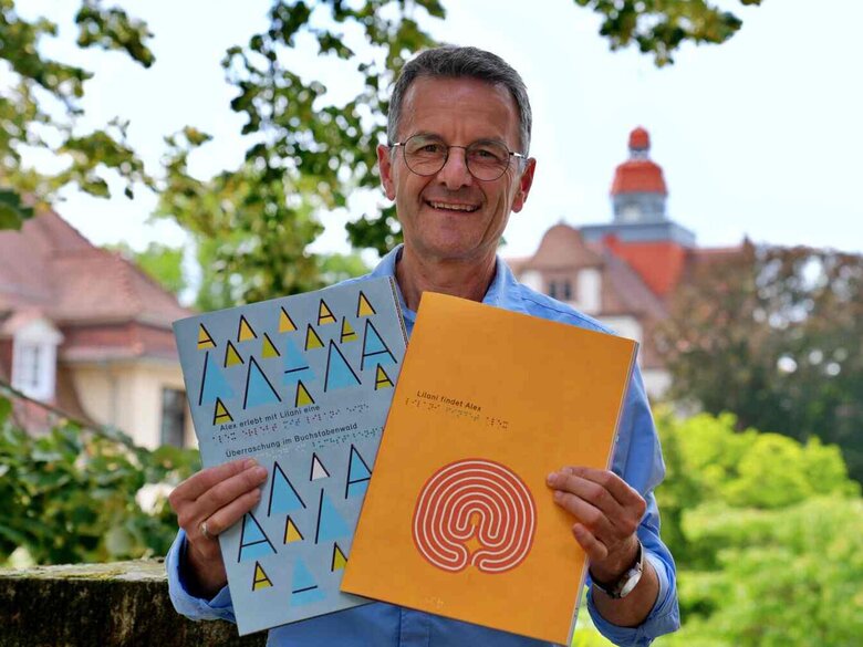 Markus Lang hält zwei Hefte Alex und Lilani in die Kamera. Er lächelt freundlich. Im Hintergrund sieht man den Altbauturm unscharf.