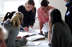 Das Bild zeigt Frauen, die sich Blätter auf einem Tisch anaschauen.