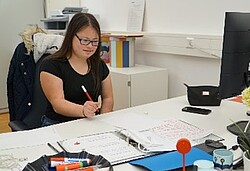 Das Foto zeigt eine junge Frau, die an einem Schreibtisch sitzt und mit dem Stift schreibt. Studierende am Tisch