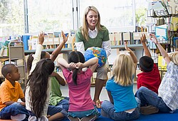 Auf dem Bild sind auf dem Boden sitzende Kinder und eine vorlesende Erzieherin.