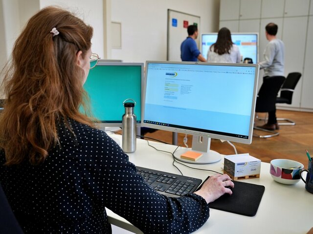 Eine Mitarbeiterin sitzt am PC. Auf dem Bildschirm sieht man eine Anmeldeseite mit dem Logo der Pädagogischen Hochschule Heidelberg. Im Hintergrund sieht man unscharf drei Personen, die an einem Stehtisch arbeiten. 