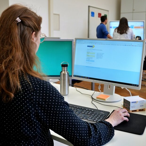 Eine Mitarbeiterin sitzt am PC. Auf dem Bildschirm sieht man eine Anmeldeseite mit dem Logo der Pädagogischen Hochschule Heidelberg. Im Hintergrund sieht man unscharf drei Personen, die an einem Stehtisch arbeiten. 