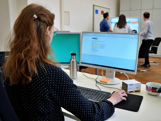 Eine Mitarbeiterin sitzt am PC. Auf dem Bildschirm sieht man eine Anmeldeseite mit dem Logo der Pädagogischen Hochschule Heidelberg. Im Hintergrund sieht man unscharf drei Personen, die an einem Stehtisch arbeiten. 
