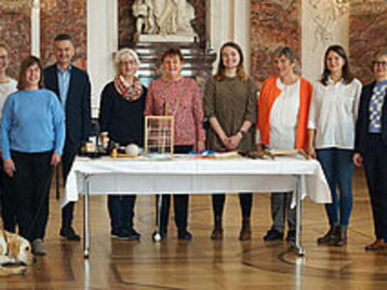 Das Foto zeigt mehrere Menschen in einer Reihe hinter einem Tisch stehen. Im Hintergrund ist die Ornamentverzierte Wand im Schloss Mannheim.