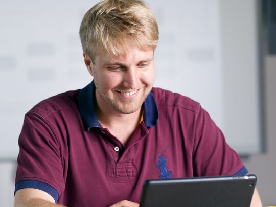 Ein junger Mann blickt lächelnd in sein Tablet. Das Bild wurde im Grünen Zimmer der Pädagogischen Hochschule Heidelberg aufgenommen