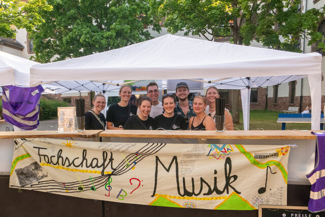 8 Studierende der Fachschaft Musik stehen bei einer Veranstaltung im Innenhof der PH hinter der Bar und lachen in die Kamera.
